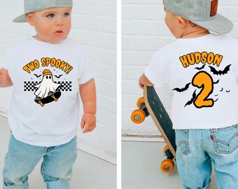 May include: White short-sleeved t-shirt with the text "TWO SPOOKY!" and a ghost riding a skateboard. The back of the shirt has the name "HUDSON" and the number "2" with bats. The child is wearing a gray baseball cap and blue jeans.