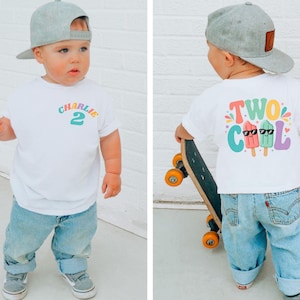 May include: White t-shirt with the name "Charlie" and the number "2" in colorful text. The back of the shirt features the words "Two Cool" with popsicle graphics wearing sunglasses. The child is wearing a gray baseball cap and light blue jeans.
