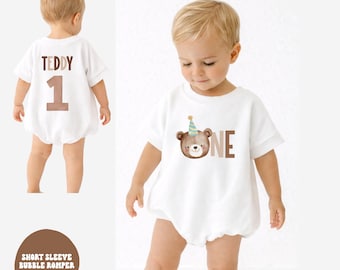 May include: White short sleeve bubble romper with a brown teddy bear graphic wearing a party hat and the word "ONE". The back of the romper has the name "TEDDY" and the number "1" in brown. The romper is for a baby's first birthday.
