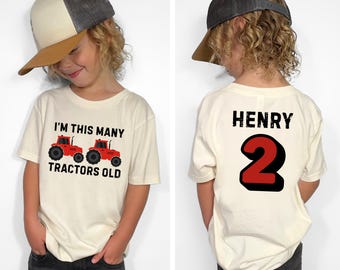 May include: A child wearing a cream-colored t-shirt and a gray and tan baseball cap. The front of the shirt says "I'M THIS MANY TRACTORS OLD" with two red tractor graphics. The back of the shirt says "HENRY" and the number "2".