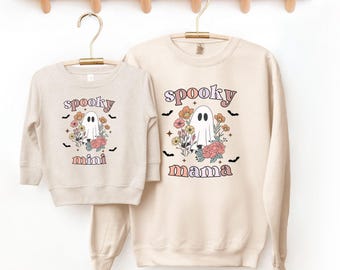 May include: Two cream colored long-sleeved shirts with a ghost graphic and floral accents. The larger shirt says "spooky mama" and the smaller shirt says "spooky mini".