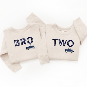 May include: Two beige sweatshirts with navy blue text and truck graphics. One sweatshirt says "BRO" and the other says "TWO". The sweatshirts are likely for a birthday or special occasion.