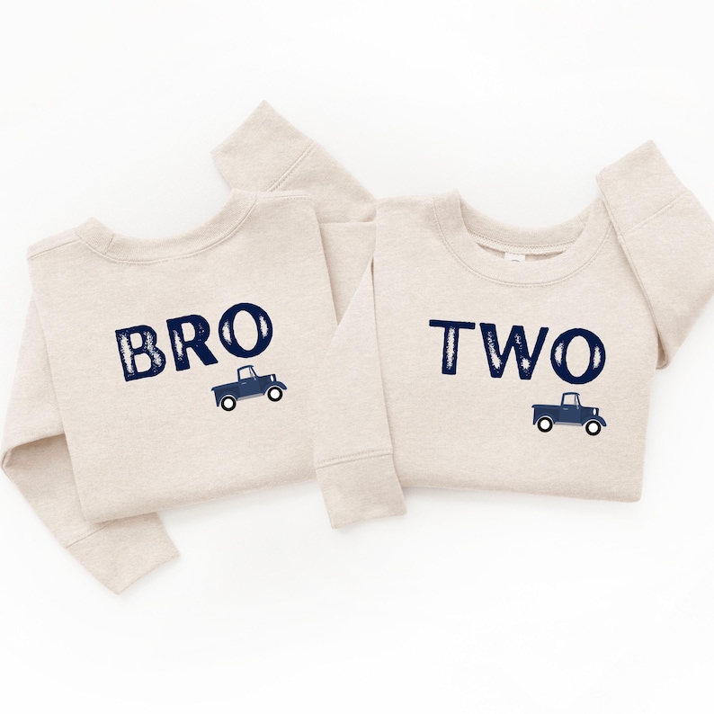 May include: Two beige sweatshirts with navy blue text and truck graphics. One sweatshirt says "BRO" and the other says "TWO". The sweatshirts are likely for a birthday or special occasion.