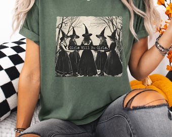May include: Dark green t-shirt featuring a vintage-style illustration of five women in witch hats and long dresses. The text "Girls Will Be Girls" is above the illustration. The shirt has rolled-up sleeves.