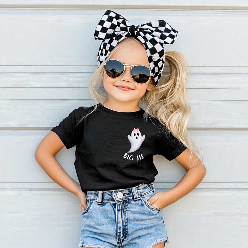 May include: A young girl wearing a black t-shirt with a ghost graphic and the words "BIG SIS". She is also wearing denim shorts, sunglasses, and a black and white checkered bow.