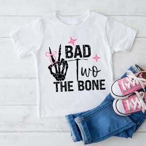 May include: White t-shirt with a black skeleton hand making a peace sign with a pink bow on the index finger. The text "Bad Two The Bone" is printed on the shirt.