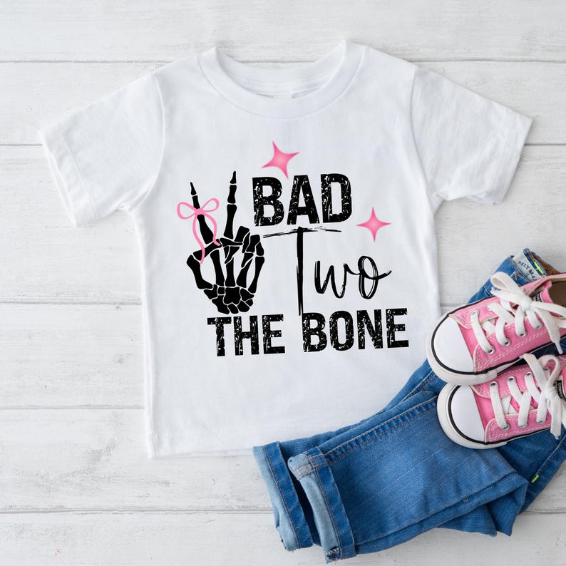 May include: White t-shirt with a black skeleton hand making a peace sign with a pink bow on the index finger. The text "Bad Two The Bone" is printed on the shirt.
