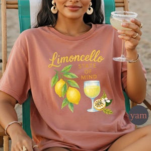 May include: A dusty rose t-shirt with the words "Limoncello State of Mind" in yellow script. The shirt features a watercolor illustration of lemons, leaves, and a glass of limoncello. A person is holding a cocktail.
