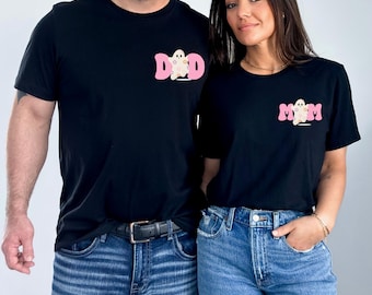 Puede incluir: Una pareja lleva camisetas negras con diseños de fantasmas rosas. La camiseta del hombre dice "DAD" y la camiseta de la mujer dice "MOM".