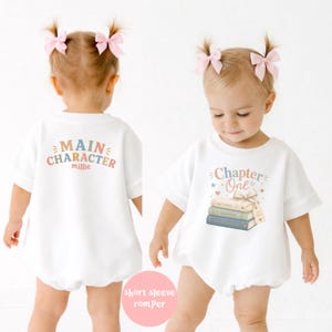 May include: A white short-sleeve romper featuring a graphic of stacked books with the text Chapter One on the front and the text Main Character Millie on the back. The design includes pastel-colored lettering, stars, and a ribbon-tied book stack.