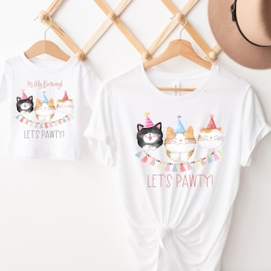 May include: Two white t-shirts with a cat birthday party theme. The larger shirt says "It's My Birthday! Let's Pawty!" and features three cats wearing party hats. The smaller shirt says "Let's Pawty!" and features three cats wearing party hats.