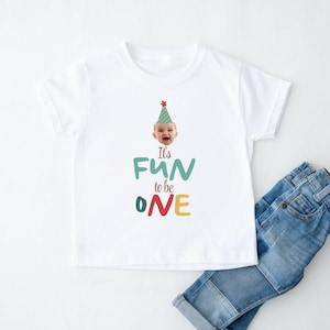 May include: White t-shirt with the text "It's FUN to be ONE" in teal, green, red, and yellow. The shirt features a smiling baby wearing a party hat. Blue denim jeans are folded next to the shirt.