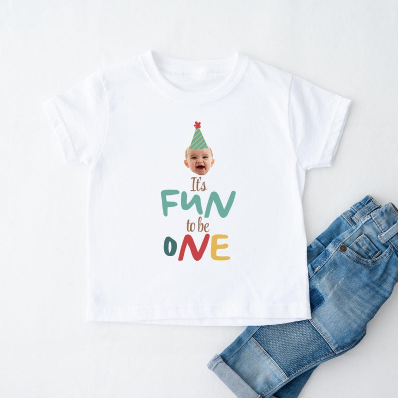 May include: White t-shirt with the text "It's FUN to be ONE" in teal, green, red, and yellow. The shirt features a smiling baby wearing a party hat. Blue denim jeans are folded next to the shirt.