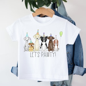 Op de afbeelding: Wit T-shirt met een afbeelding van zes honden die feestmutsen dragen en de tekst "Let's Pawty!"