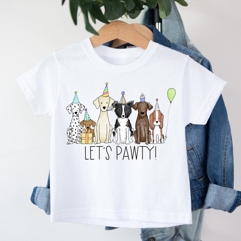 Op de afbeelding: Wit T-shirt met een afbeelding van zes honden die feestmutsen dragen en de tekst "Let's Pawty!"