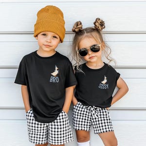 May include: Two children wearing matching black t-shirts with a cartoon duck design and the words "BRO" and "SISSY". They are also wearing black and white checkered shorts and a mustard yellow beanie. One child is wearing sunglasses.