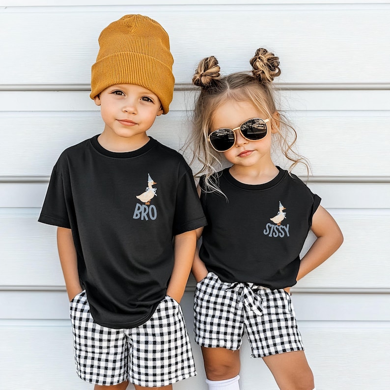 May include: Two children wearing matching black t-shirts with a cartoon duck design and the words "BRO" and "SISSY". They are also wearing black and white checkered shorts and a mustard yellow beanie. One child is wearing sunglasses.