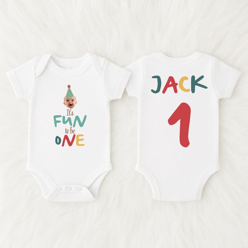 May include: White baby bodysuit with the text "It's FUN to be ONE" in teal, green, red, and yellow. The front of the bodysuit features a baby wearing a party hat. The back of the bodysuit says "JACK 1" in green, yellow, and red.