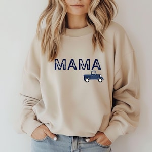 May include: Beige sweatshirt with the word "MAMA" in blue block letters and a blue truck graphic. The sweatshirt is a neutral color and has a crew neck.