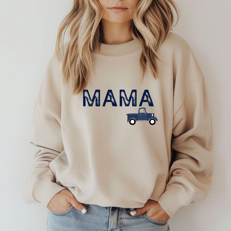 May include: Beige sweatshirt with the word "MAMA" in blue block letters and a blue truck graphic. The sweatshirt is a neutral color and has a crew neck.