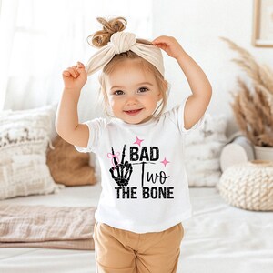 May include: A white short-sleeved t-shirt with a black skeleton hand making a peace sign. The shirt has pink glitter and the text "Bad Two The Bone".