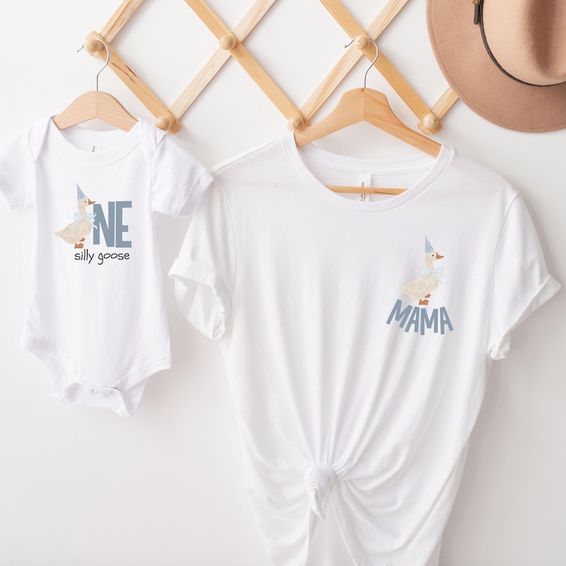 May include: White baby onesie with a cartoon goose wearing a party hat and the word "ONE" in blue. A white t-shirt with a matching goose design and the word "MAMA" in blue is also displayed.