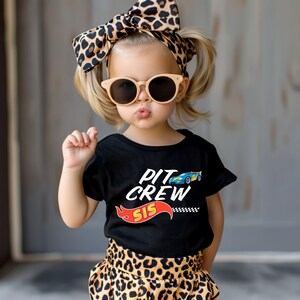 May include: A young girl wearing a black t-shirt with the text "Pit Crew Sis" and a cartoon race car. She is also wearing a leopard print headband and sunglasses.