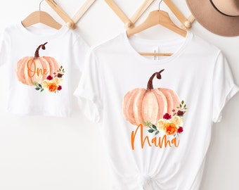 Könnte beinhalten: Zwei weiße T-Shirts mit einem Aquarellkürbis und Blumendesign. Ein Shirt sagt "Mama" und das andere "One".