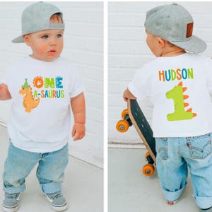 May include: A white t-shirt with a dinosaur graphic and the text "ONE A-SAURUS" on the front. The back of the shirt has the name "HUDSON" and a number "1" dinosaur graphic. The child is wearing blue jeans and a gray baseball cap.