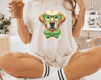 Può includere: Una maglietta bianca con una grafica di un golden retriever che indossa occhiali verdi e un papillon verde con pois bianchi. Il cane indossa un trifoglio su ogni orecchio.