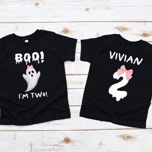 May include: Two black t-shirts with white text and graphics. One shirt says "BOO! I'M TWO!" with a ghost graphic and pink bow. The other shirt says "VIVIAN" and has a number "2" with a pink bow. The shirts are on a light wood surface.