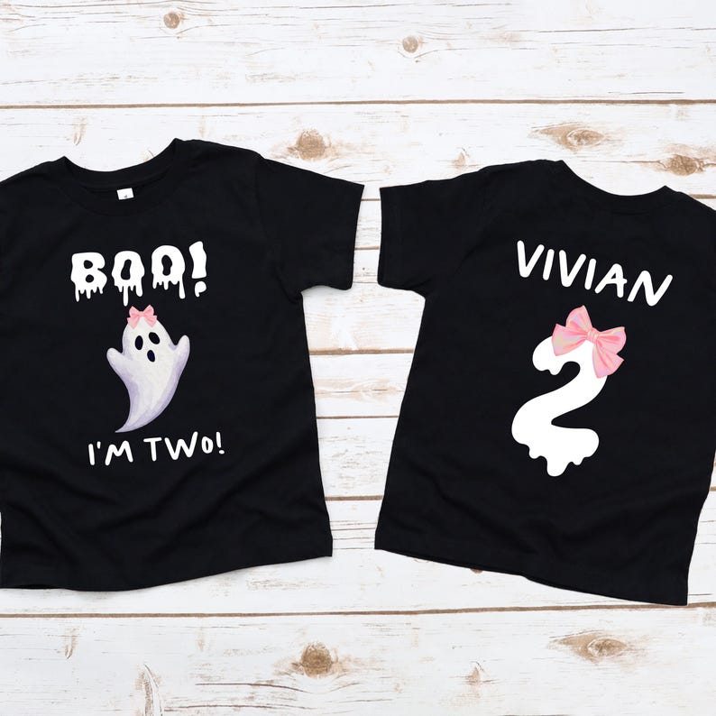 May include: Two black t-shirts with white text and graphics. One shirt says "BOO! I'M TWO!" with a ghost graphic and pink bow. The other shirt says "VIVIAN" and has a number "2" with a pink bow. The shirts are on a light wood surface.