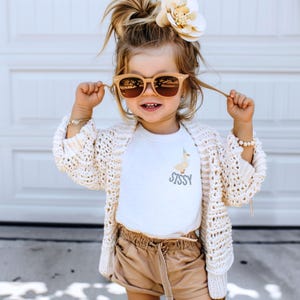 May include: A young child wearing sunglasses, a white t-shirt with a duck graphic and the word "Sissy", a cream-colored crochet cardigan, and tan shorts. A large flower is in the child's hair.