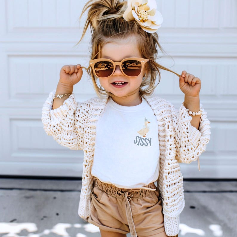 May include: A young child wearing sunglasses, a white t-shirt with a duck graphic and the word "Sissy", a cream-colored crochet cardigan, and tan shorts. A large flower is in the child's hair.