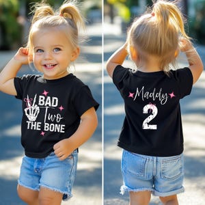 May include: A black toddler t-shirt with the text "BAD TWO THE BONE" in white and pink. The back of the shirt has the name "Maddy" and the number "2" in white. The toddler is wearing blue denim shorts.