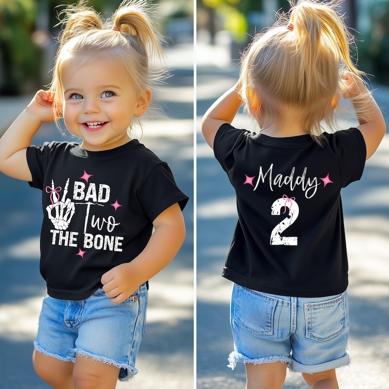 May include: Black t-shirt with white text that says "I Bad Two The Bone" with a pink bow and a skeleton hand making a peace sign. The back of the shirt has white text that says "Maddy" with a pink bow and the number "2".