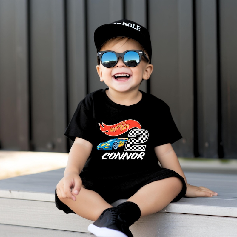 May include: A black t-shirt with a red and yellow race car graphic and the text "Happy Birthday Boy 2 CONNOR". A young child is wearing the shirt and sunglasses.
