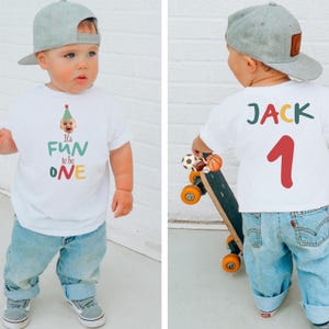 May include: A toddler wearing a white t-shirt with the text "It's FUN to be ONE" and a birthday hat graphic. The child is also wearing blue jeans and a gray baseball cap. The back of the shirt says "JACK 1".
