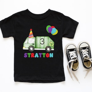 May include: Black t-shirt with a green garbage truck with a party hat and balloons. The truck has a recycle symbol and the number 3 on the side. The text "STRATTON" is printed below the truck.