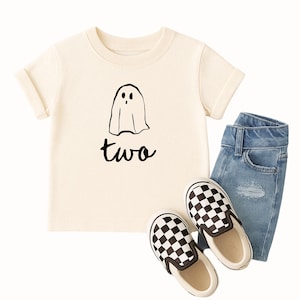 May include: A cream-coloured t-shirt featuring a black ghost illustration and the word "two". The outfit includes light blue denim jeans and black and white chequered slip-on shoes. A casual outfit for a toddler.