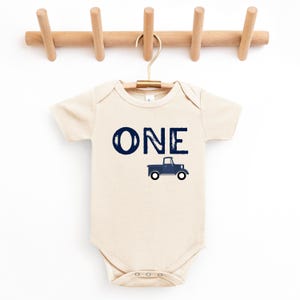 May include: A cream-colored baby onesie with the word "ONE" in navy blue script and a navy blue truck graphic. The onesie is hanging on a wooden hanger.