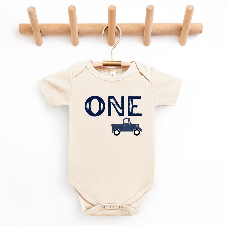May include: A cream-colored baby onesie with the word "ONE" in navy blue script and a navy blue truck graphic. The onesie is hanging on a wooden hanger.