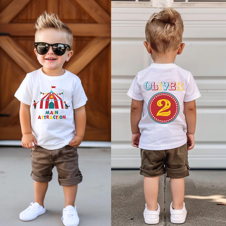 May include: A young child wearing a white t-shirt with a circus-themed graphic and the words "MAIN ATTRACTION." The child is also wearing brown shorts, white sneakers, and sunglasses. The back of the shirt has the name "OLIVER" and the number "2."