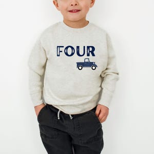 May include: A light gray sweatshirt with the word "FOUR" in navy blue block letters and a navy blue truck graphic. The sweatshirt is worn by a child, paired with black pants.