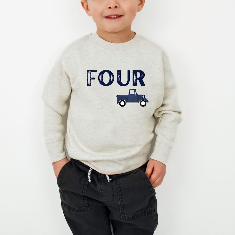 May include: A light gray sweatshirt with the word "FOUR" in navy blue block letters and a navy blue truck graphic. The sweatshirt is worn by a child, paired with black pants.