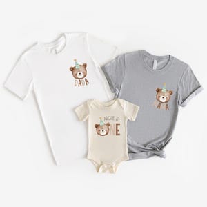 May include: A set of three shirts featuring a brown bear wearing a party hat. The shirts are white, gray, and beige. The white shirt says "Dada", the gray shirt says "Mama", and the beige shirt says "Archie is One".