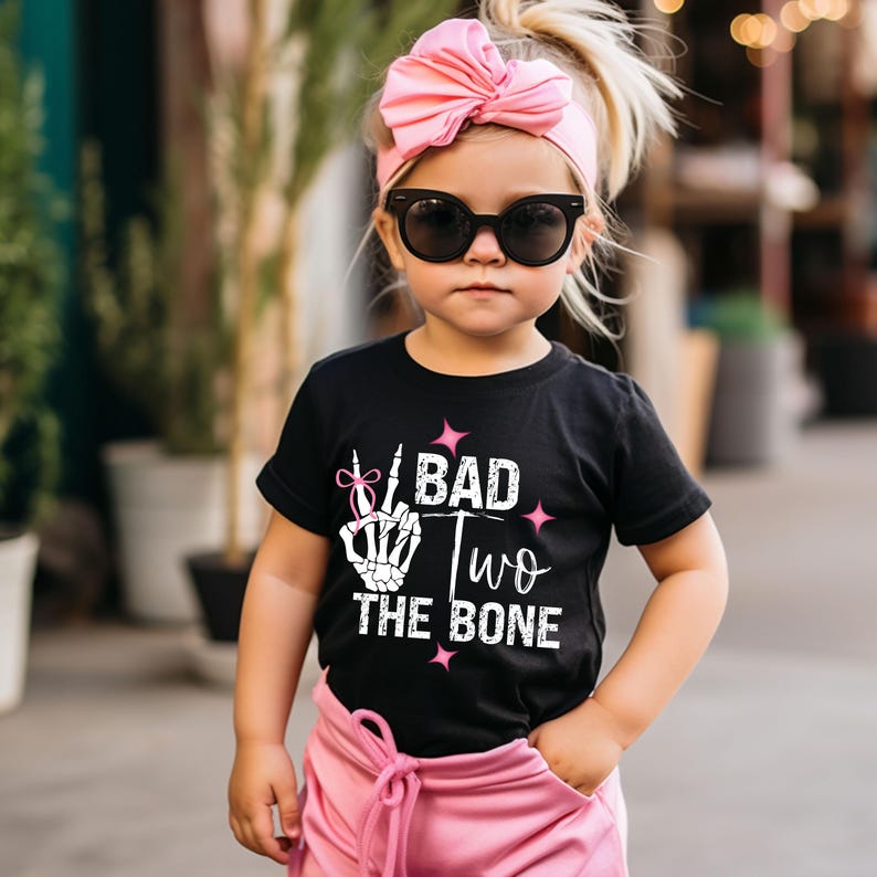 May include: A black t-shirt with a white skeleton hand making a peace sign and the text "I BAD TWO THE BONE". The shirt has pink glitter accents. A young child is wearing the shirt and a pink headband.