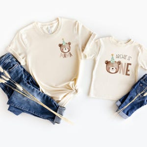 May include: Two beige t-shirts with a brown bear wearing a party hat. The larger shirt says "Mama" and the smaller shirt says "Archie is One".