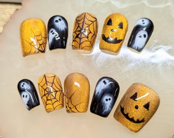 May include: A set of ten Halloween-themed false nails. Designs include gold glitter, black spiderwebs, white ghosts, and jack-o'-lantern faces. The nails are arranged on a light-colored, oval-shaped surface.