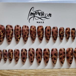 Könnte beinhalten: Set mit 20 falschen Nägeln mit einem braunen und goldenen Leopardenmuster. Die Nägel sind in zwei Reihen auf einer weißen Karte mit dem Text "Impress On Nails" angeordnet.